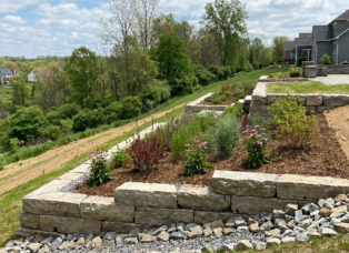 A professionally engineered multi-tiered limestone retaining wall stabilizing a steep backyard slope in Clarence, NY, featuring native deep-rooted plantings and protective coir matting to prevent soil erosion.