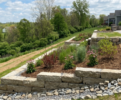 A professionally engineered multi-tiered limestone retaining wall stabilizing a steep backyard slope in Clarence, NY, featuring native deep-rooted plantings and protective coir matting to prevent soil erosion.