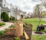 A pristine suburban lawn in Clarence NY showing a garden bed clean up with fresh mulch, a paper leaf bag, straw broom, and garden utility cart during spring landscaping preparation.